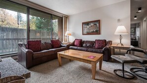 A living room with a brown leather sofa, a wooden coffee table, and a grey armchair. There is a painting on the wall and a lamp on a side table.