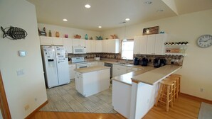 A modern kitchen with white cabinetry, a central island with stools, a microwave, a sink, and a clock on the wall. - KH4004- Oceanside-5 BRs, Elevator, Pool Table, Private Pool and Hot Tub! (Kitty Hawk)