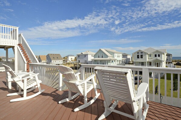 A wooden deck with white rocking chairs and a white railing overlooking a residential area with houses and a clear sky. - KH4004- Oceanside-5 BRs, Elevator, Pool Table, Private Pool and Hot Tub! (Kitty Hawk)