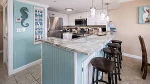 Una cocina con pared de acento color aguamarina, gabinetes blancos, refrigerador de acero inoxidable, microondas y encimera de granito. Hay cuatro taburetes de bar y una lámpara colgante.