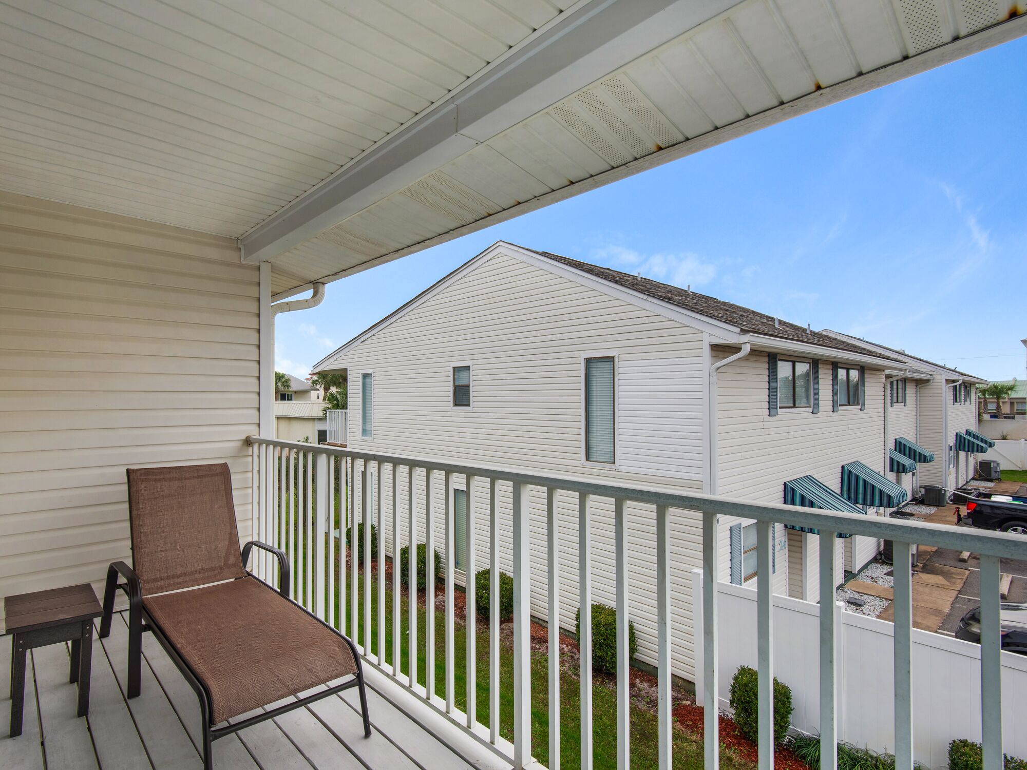 A balcony with a chair and a small table, overlooking a white house with a green roof and a white fence.
