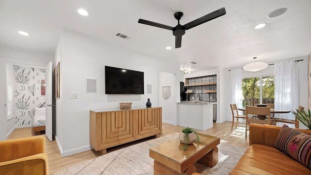 A modern living room with a sofa, coffee table, and TV. There is a ceiling fan, a kitchen area, and a dining table with chairs. - Designer Duplex | Pool, Fire Pit, Near UT Football (Austin)