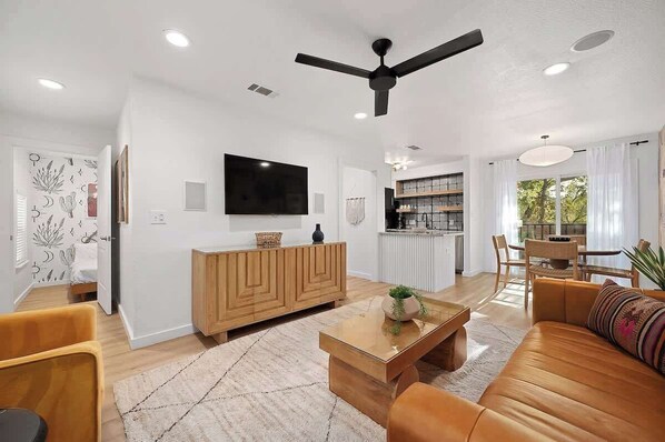 A modern living room with a sofa, coffee table, and TV. There is a ceiling fan, a kitchen area, and a dining table with chairs. - Designer Duplex | Pool, Fire Pit, Near UT Football (Austin)