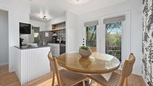 A modern kitchen with white cabinetry, a central island, and a dining area with a glass table and woven chairs. - Designer Duplex | Pool, Fire Pit, Near UT Football (Austin)
