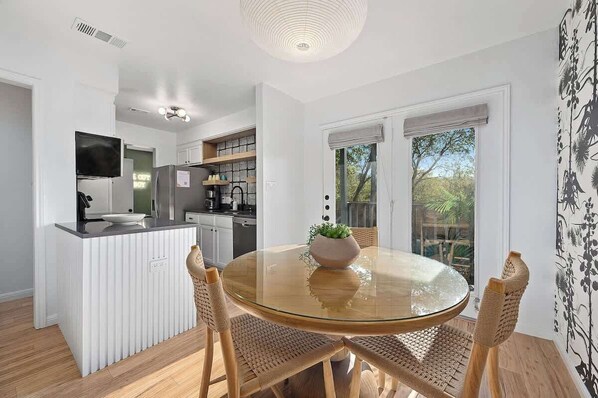 A modern kitchen with white cabinetry, a central island, and a dining area with a glass table and woven chairs. - Designer Duplex | Pool, Fire Pit, Near UT Football (Austin)