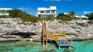 Une propriété située sur la côte, avec un escalier en bois menant à une terrasse surplombant l’eau, une eau turquoise et limpide, et un bâtiment blanc en arrière-plan.