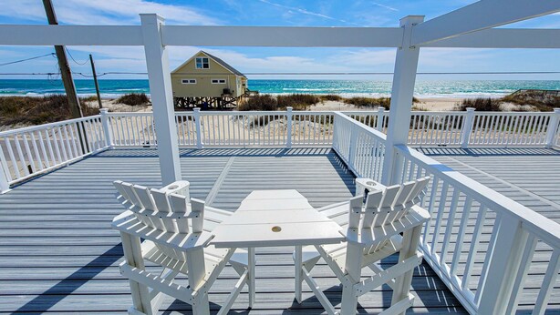 A wooden deck with white railings and a table and chairs, overlooking a beach and a house. - Unwind in Style at Elysium (Surf City)