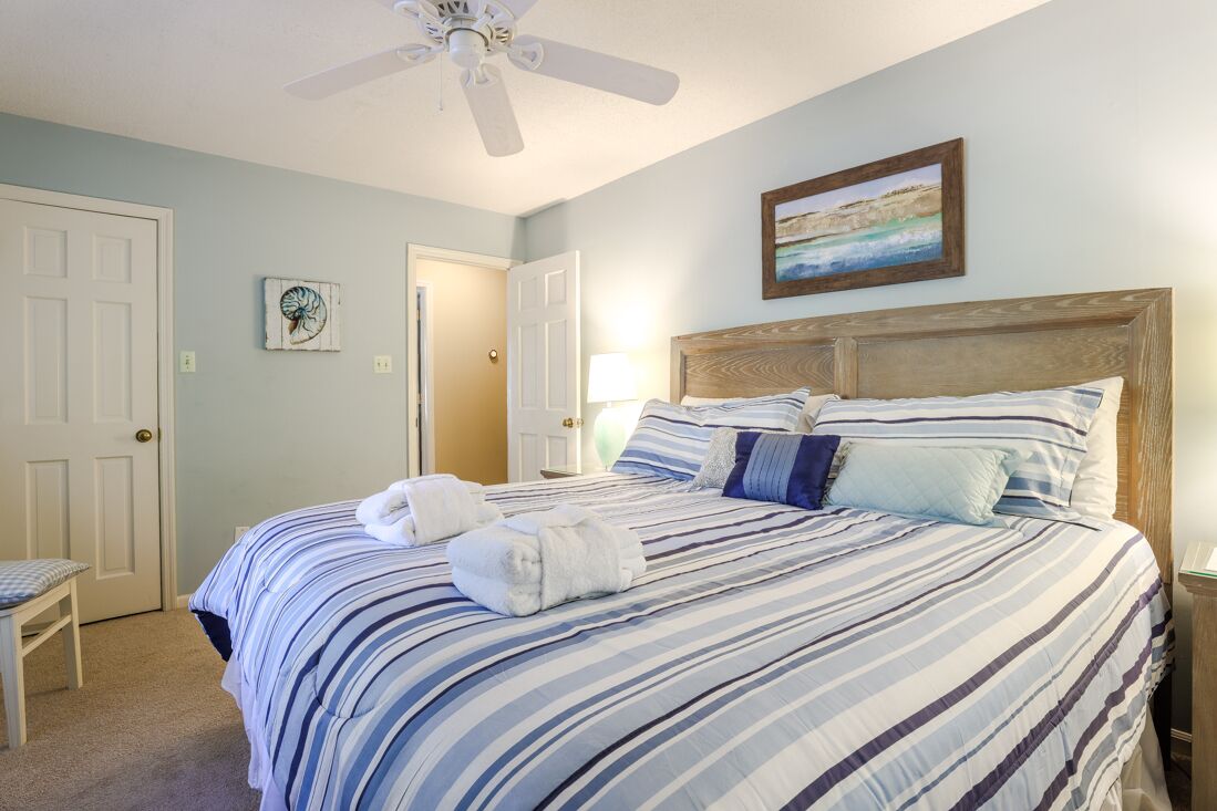 A neatly made bed with striped bedding, a ceiling fan, and a painting on the wall.