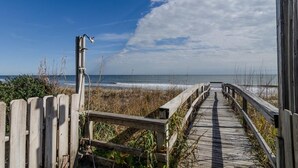 Ein hölzerner Steg führt zum Strand, im Hintergrund ist ein klarer Himmel und der Ozean zu sehen.