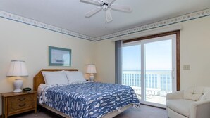 Ein Schlafzimmer mit einem Bett, zwei Nachttischlampen, einem Nachttisch, einem gerahmten Bild, einem Deckenventilator und einem Balkon mit Meerblick.