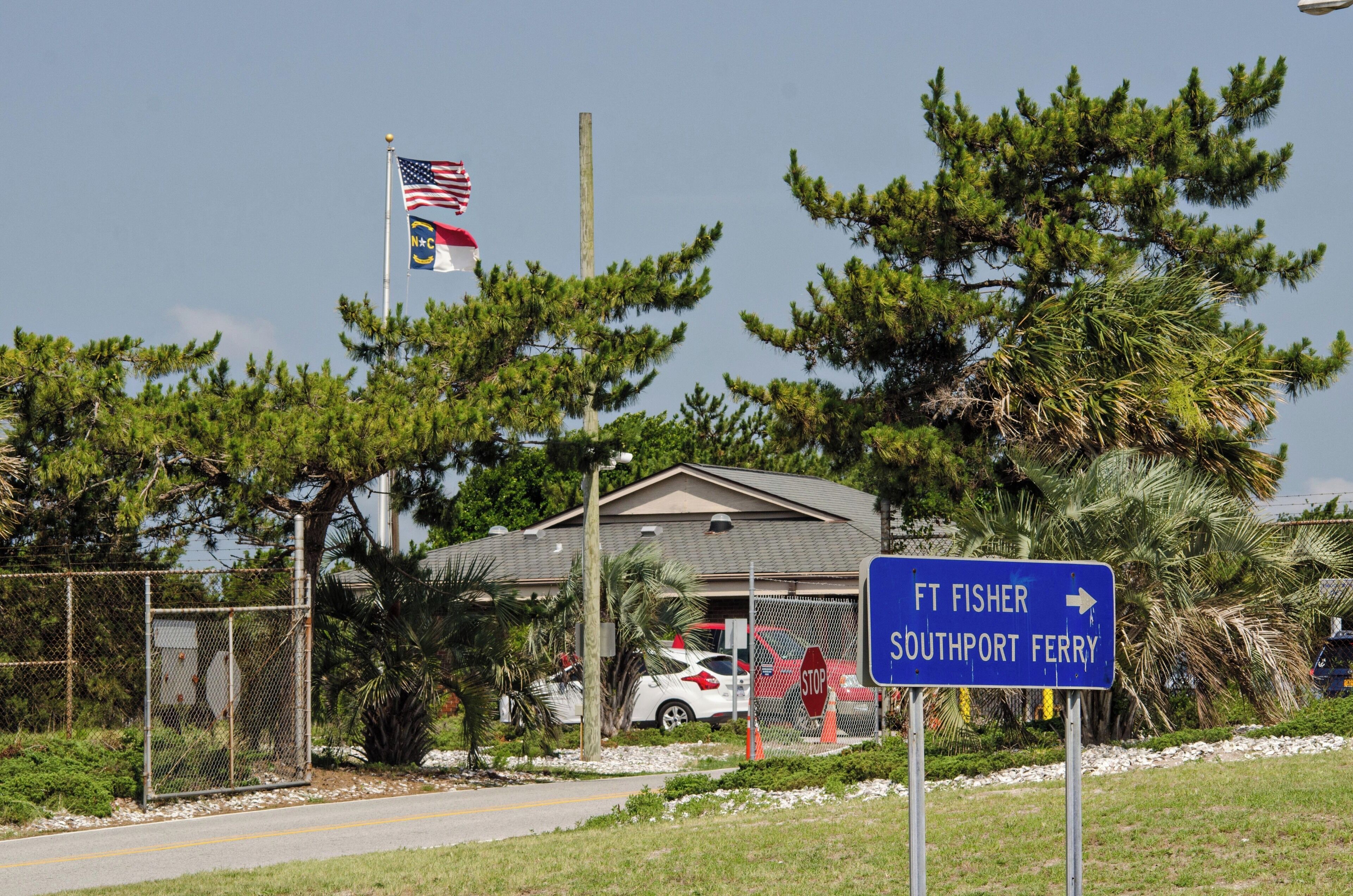 A sign for Ft. Fisher Southport Ferry, a fenced area with a building, cars, and a flag.