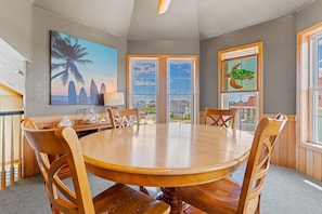 A dining area with a round table and wooden chairs, large windows with a view, and artwork on the walls.