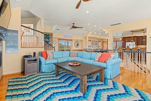 A spacious living room with a large L-shaped sectional sofa, a wooden coffee table, and a patterned rug. A staircase with a railing leads to an upper level.