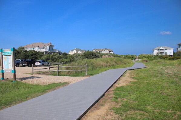 A boardwalk leading to beachfront houses with a sign indicating public beach parking. - Marguerite DeVille, Custom-designed Oceanside Home, All Seasons Getaway (Corolla)