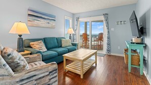 Ein Wohnzimmer mit einer Couch, einem Couchtisch und einem Fernseher auf einem Schrank. Es gibt einen Balkon mit Stühlen und Blick aufs Meer.