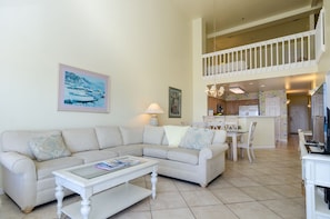 A living room with a sofa, coffee table, and a dining area with chairs and a table. There is a kitchen in the background with cabinets and a refrigerator.