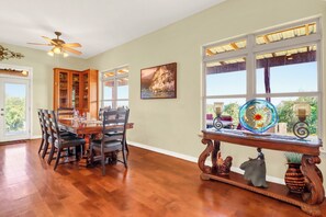 Un coin repas avec une table et des chaises en bois, un ventilateur de plafond et une fenêtre donnant sur un paysage verdoyant.