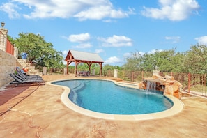 A swimming pool with a waterfall feature, a covered seating area, and a stone wall.