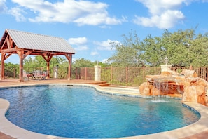 A swimming pool with a waterfall feature, a covered pavilion, and a fenced area with trees.