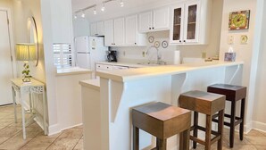 A modern kitchen with white cabinetry, a central island with stools, a coffee maker, and a sink. - Direct Ocean & Beach View -Litchfield, Rare 2 Oceanfront Porches! (Pawleys Island)