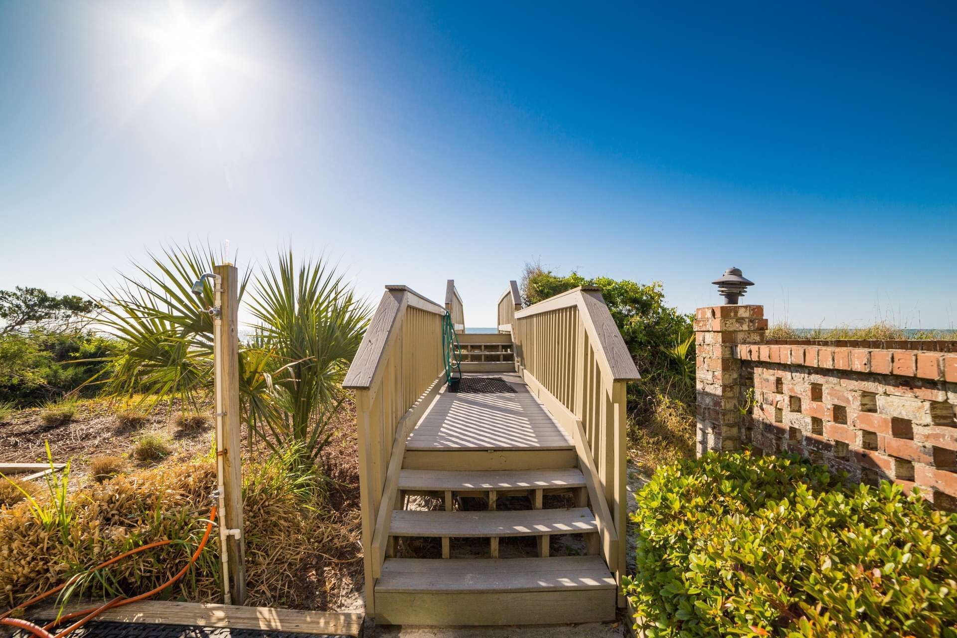 A wooden staircase leading to a beach area with a clear sky and a brick wall on the right.
