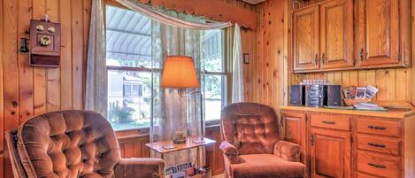 A cozy room with wooden walls and a large window with curtains. There are two plush armchairs, a small table with a lamp, and a cabinet with kitchen appliances.