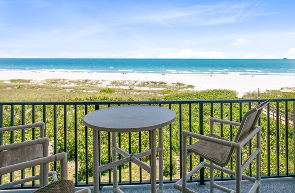 Un balcon avec une table et deux chaises, donnant sur une plage de sable blanc et une eau d’un bleu cristallin.