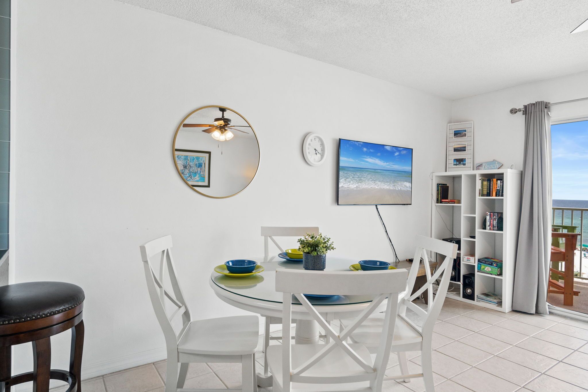 A dining area with a round table, white chairs, a ceiling fan, a clock, a bookshelf, and a view of the sea.