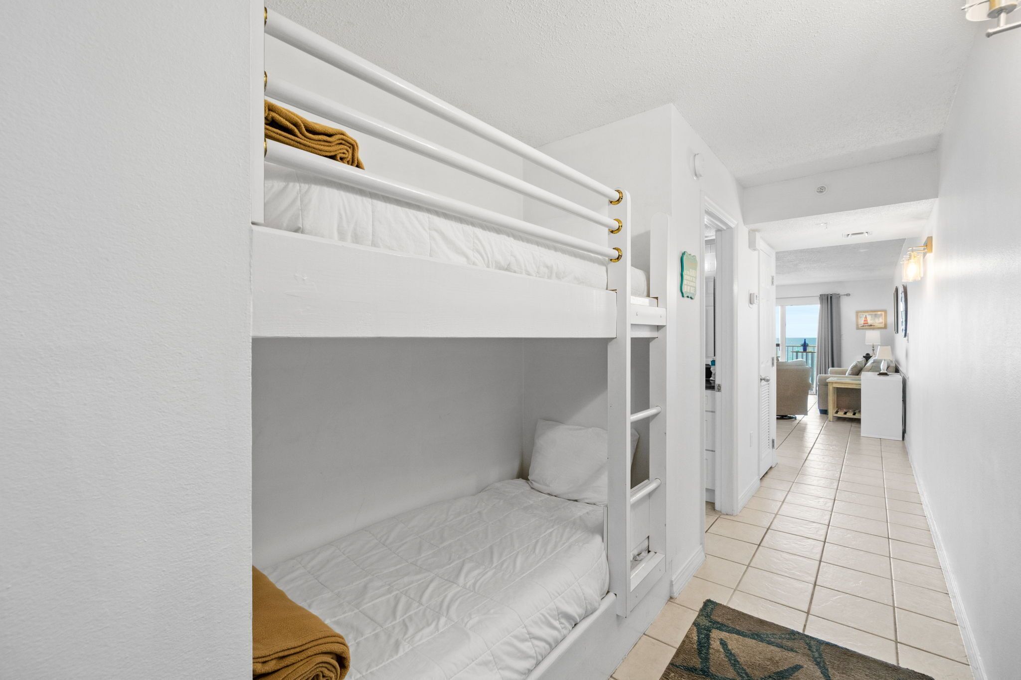 Dormitorio con literas, una paleta de colores blancos, un pasillo que conduce a un baño y una pequeña zona de estar con una mesa y sillas.