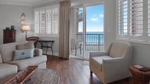Sala de estar com sofá, poltrona e mesa pequena. Vista para o oceano através de grandes janelas.