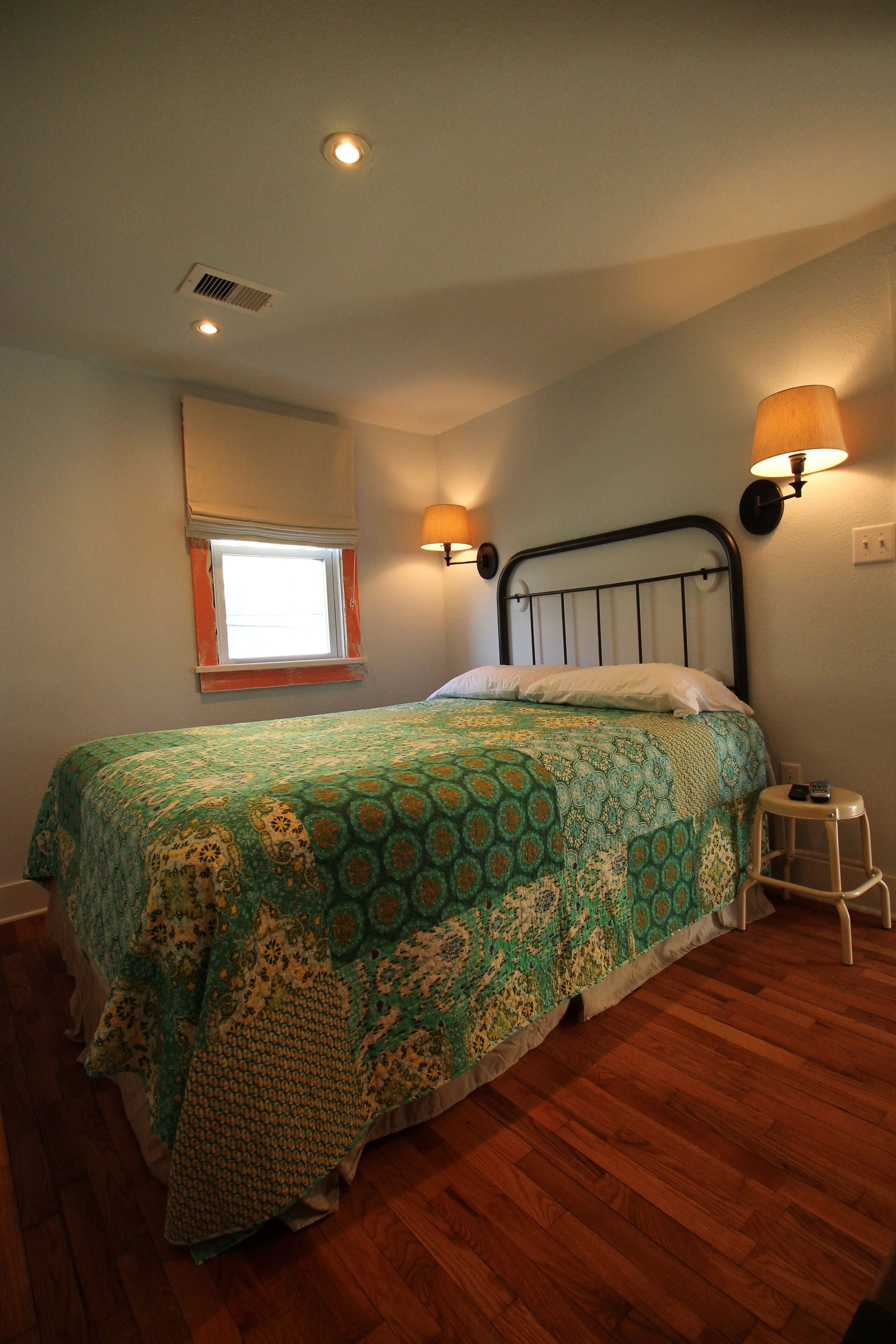 A bedroom with a bed, a window, a wall-mounted lamp, and a wooden floor.