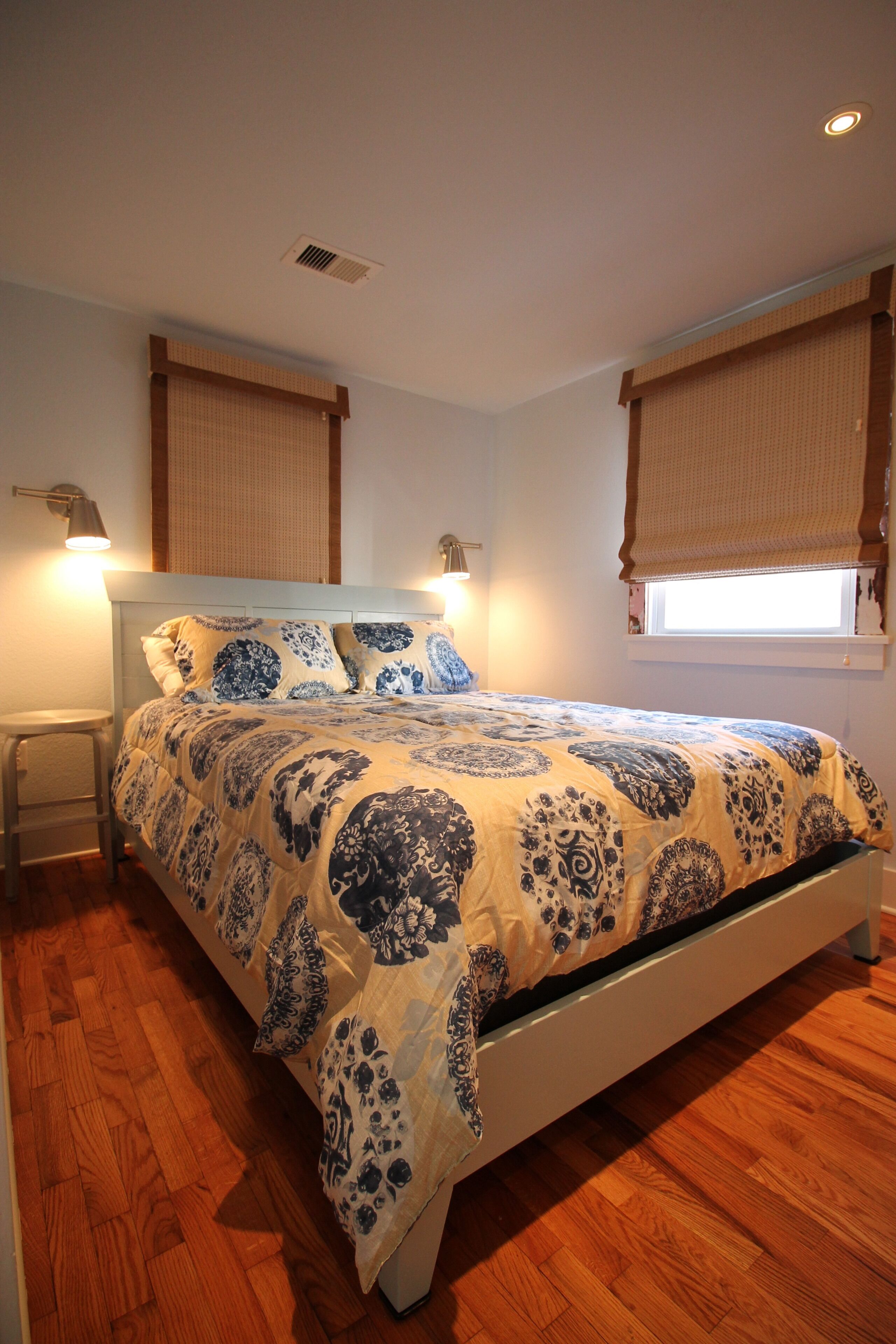 A bedroom with a bed, wooden flooring, two windows with blinds, and a nightstand.
