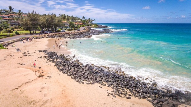 A beach with rocky shoreline, clear turquoise water, and a residential area with houses in the background. - Luxury Poipu 4br Villa with AC Walk to the Beach! (224) (Koloa)