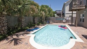 A rectangular swimming pool with a curved edge, surrounded by a brick patio. There are palm trees and a wooden fence in the background.