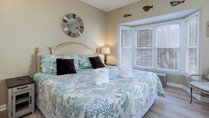 Une chambre à coucher comprenant un lit, une table de chevet, une lampe, une fenêtre avec des stores et des éléments de décoration murale.