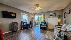 A living room with a ceiling fan, a TV, a dining area with chairs, a sofa, and a view through a glass door to the outside. - Stay Coastal at Fenway at the Beach (North Topsail Beach)