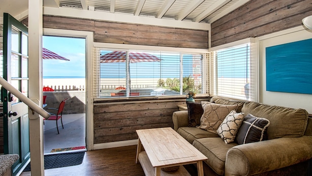 Een gezellige kamer met een houten plafond, een raam met uitzicht op het strand, een bank met gestreepte kussens en een klein houten tafeltje.