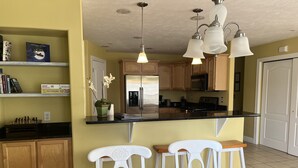 Una cocina con armarios de madera, un refrigerador, un microondas y una estufa. Hay una mesa de comedor con sillas y una lámpara colgante arriba.