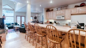 A modern kitchen with wooden bar stools, a dining area with a chandelier, and a cozy living room with a large window.