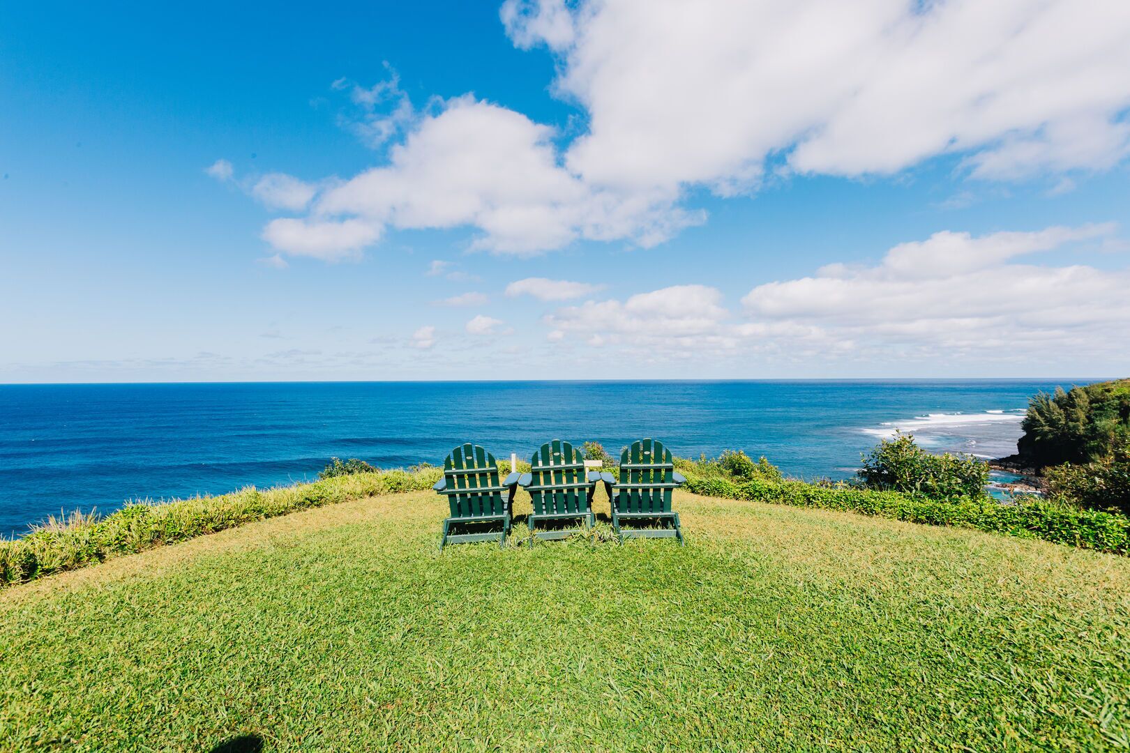 A grassy area with three green Adirondack chairs facing a view of the ocean.