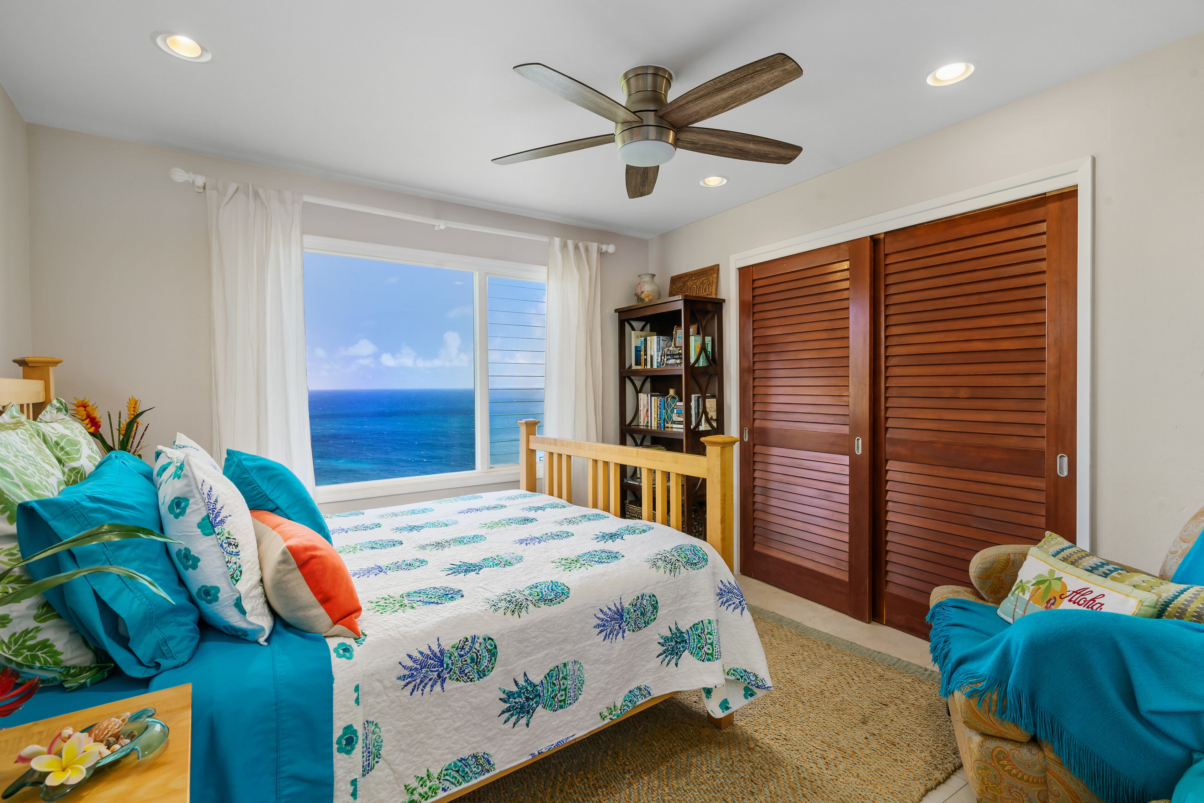 A bedroom with a bed, a wooden chair, a bookshelf, and a view of the ocean.