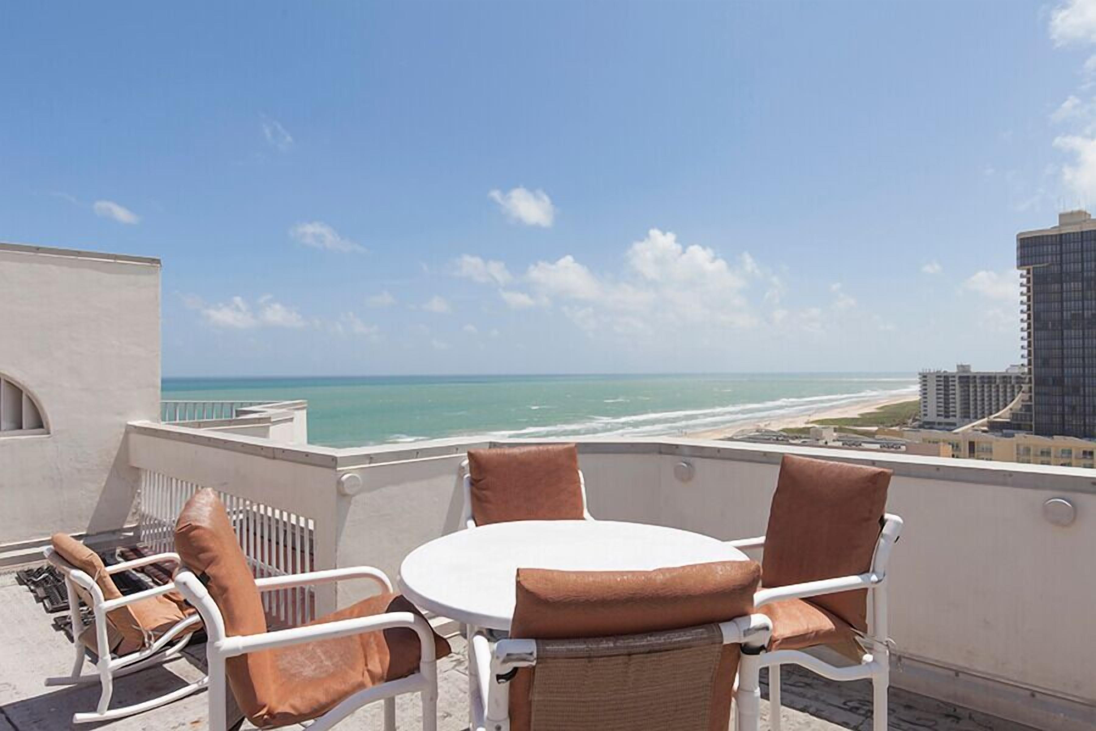 A balcony with a white table and chairs, overlooking a beach and ocean.