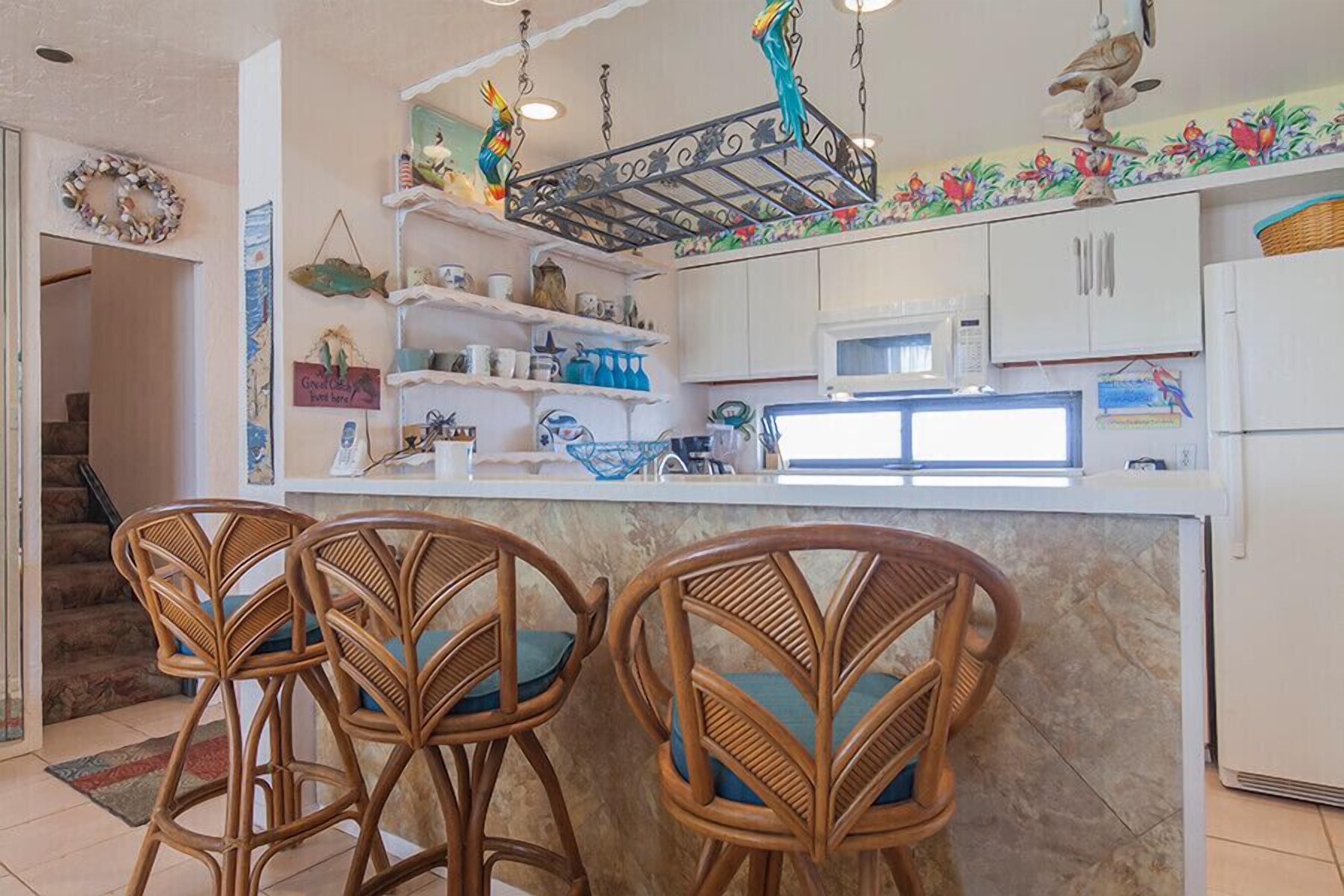A kitchen with a marble countertop, white cabinets, a microwave, and a hanging light fixture. There are two wicker chairs with teal cushions.
