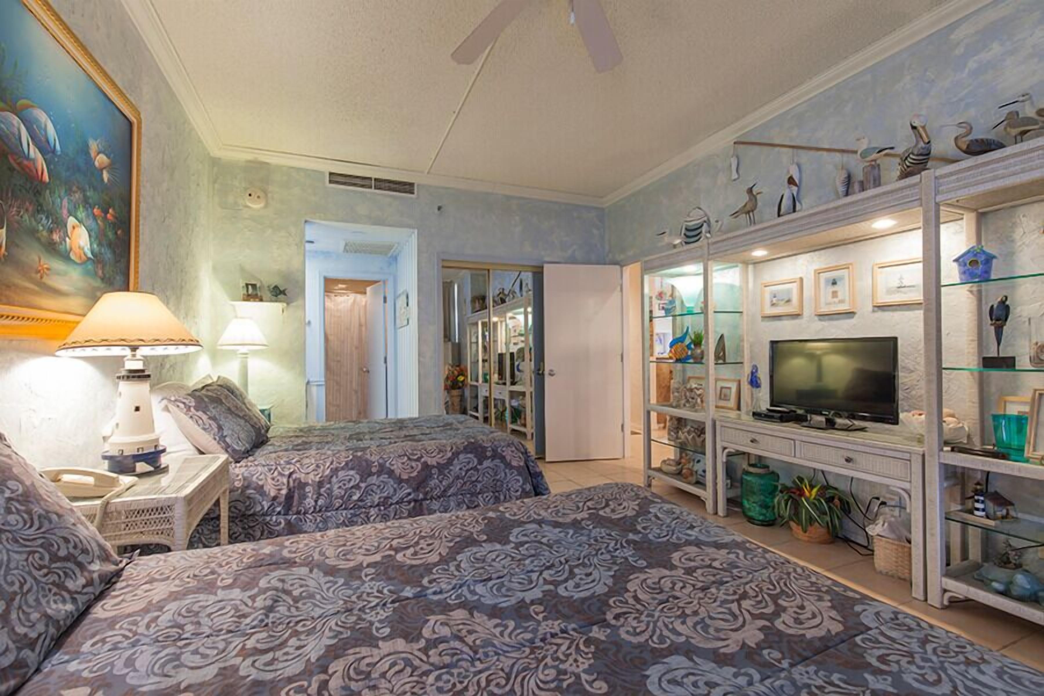 A hotel room with two beds, a television, a glass display cabinet with decorative items, and a painting of fish on the wall.
