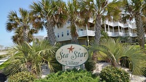 A sign for Sea Star Village with a star emblem, surrounded by palm trees and a building with balconies. - Anchored Inn: Family-Friendly 4BR with Pool (Surf City)