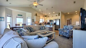 A spacious living room with a sofa, coffee table, and a patterned armchair. A ceiling fan and pendant lights are visible. - Anchored Inn: Family-Friendly 4BR with Pool (Surf City)