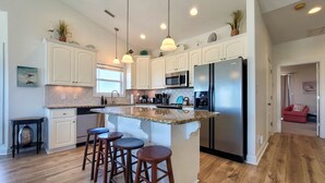 A modern kitchen with white cabinets, a central island with stools, stainless steel appliances, and pendant lighting. - Anchored Inn: Family-Friendly 4BR with Pool (Surf City)