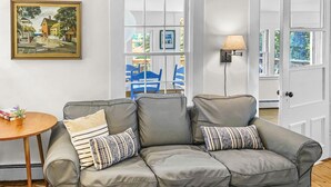 Ein graues Ecksofa mit zwei gemusterten Kissen, ein kleiner Holztisch mit einem Buch und einer Pflanze, ein gerahmtes Landschaftsbild und eine Lampe auf einer Wandhalterung.
