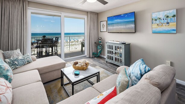 A living room with a large window overlooking the beach, a ceiling fan, and a flat-screen TV on the wall.