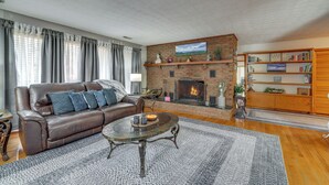 Una sala de estar con un sofá de cuero, una chimenea, una estantería y un amplio ventanal con cortinas.
